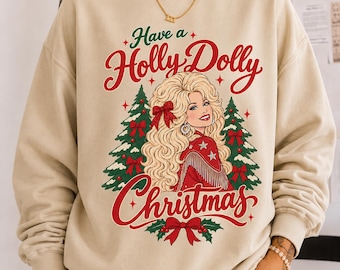 May include: A beige crewneck sweatshirt featuring a graphic print of a blonde woman with voluminous hair, wearing a red fringed jacket. The text reads Have a Holly Dolly Christmas, surrounded by green Christmas trees, red bows, and stars.
