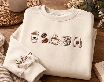 May include: Cream-colored sweatshirt with embroidered coffee-themed icons: a to-go cup with a heart, coffee beans, a steaming cup, a coffee machine, and a bag of beans. The sleeve cuff features the name "Sofia" embroidered in brown.