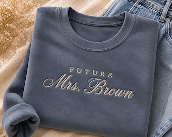 May include: A slate blue crewneck sweatshirt featuring the embroidered text FUTURE Mrs. Brown in a cream-colored thread. The garment has a ribbed collar and long sleeves, with the word FUTURE in a sans-serif font above the cursive script Mrs. Brown.