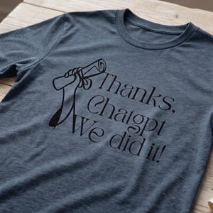 May include: A blue t-shirt with the text "Thanks, Chatgpt, We did it!" in black script. The shirt features a graduation cap and diploma graphic. The shirt is displayed on a wooden surface.