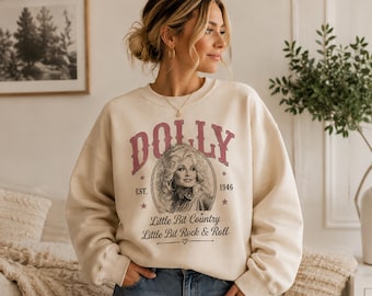 May include: A cream-colored crewneck sweatshirt featuring a graphic print of Dolly Parton in a circular frame. The text reads DOLLY, EST. 1946, Little Bit Country, and Little Bit Rock & Roll in red and black lettering with small star accents.