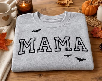 May include: A light gray crewneck sweatshirt featuring the word MAMA embroidered in black block letters. The letters include a spiderweb pattern detail. Three small black bats are embroidered around the text, with one above the M and two below the A.