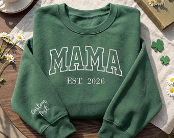 May include: A forest green sweatshirt with the word "MAMA" in white block letters, with "EST. 2026" below. The sleeve reads "Custom Text" in white script. The sweatshirt is on a wooden surface with flowers.