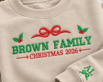 May include: A beige crewneck sweatshirt featuring embroidered text that reads BROWN FAMILY CHRISTMAS 2026. The design includes a red bow, green holly leaves with red berries, and the initials AL embroidered in green on the sleeve cuff.