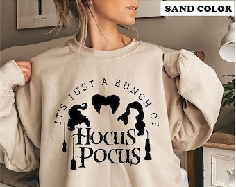 May include: A sand-colored crewneck sweatshirt featuring a black graphic print. The design includes the text 'IT'S JUST A BUNCH OF HOCUS POCUS' arranged around three stylized silhouettes of hair and two broomstick icons centered on the chest.