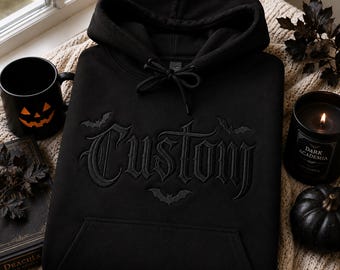 May include: A black hooded sweatshirt featuring the word Custom embroidered in a gothic font across the chest, accented by three small embroidered bat silhouettes. The garment includes a front kangaroo pocket and matching black drawstring cords.