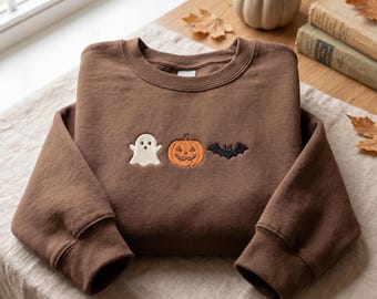 May include: A brown crewneck sweatshirt featuring an embroidered design on the chest. The embroidery consists of a white ghost, an orange jack-o'-lantern pumpkin, and a black bat arranged in a horizontal row across the center of the garment.