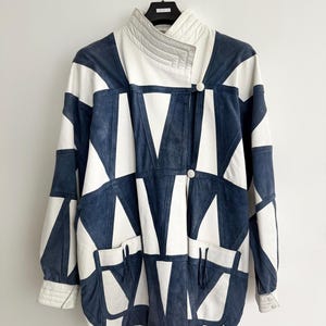 May include: A vintage jacket in navy blue and white with a bold geometric design. The jacket has a high collar, two front pockets, and button fastenings. The cuffs of the sleeves have a quilted detail. It is displayed on a metal hanger.