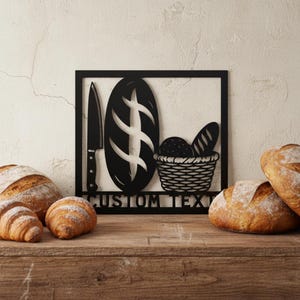 Könnte beinhalten: Schwarze Metall-Wandkunst mit einem Brotlaib, einem Messer und einem Brotkorb. Die Worte "CUSTOM TEXT" stehen unter dem Brot und dem Korb. Frisch gebackenes Brot und Croissants liegen auf einer Holzoberfläche.