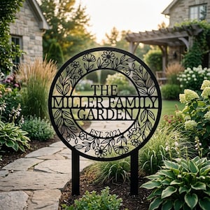 Könnte beinhalten: Ein kreisförmiges schwarzes Metall-Gartenschild mit einem floralen Kranzrand und dem Text THE MILLER FAMILY GARDEN EST. 2021. Das Schild wird von zwei vertikalen Pfosten gestützt und befindet sich in einem Garten mit Pflanzen und Steinwegen.