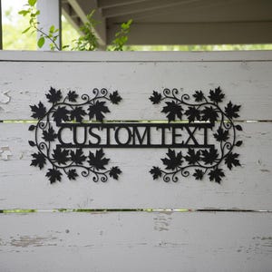 Könnte beinhalten: Schwarzes Metallschild mit dem Text "CUSTOM TEXT" in einem rechteckigen Rahmen. Der Rahmen ist mit Ahornblattmotiven und dekorativen Wirbeln verziert. Das Schild ist an einem weißen Holzzaun befestigt.