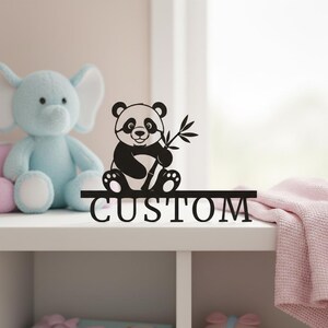Peut inclure: Décoration en forme de panda noir et blanc tenant du bambou, avec le mot « CUSTOM » découpé. Posé sur une étagère blanche, avec un éléphant bleu et une couverture rose en arrière-plan.