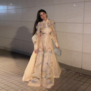 May include: A floor-length, long-sleeved dress in a pale peach hue, featuring a high neckline and a delicate, intricate floral pattern in shades of blue, green, and gold. The garment has a fitted bodice, a cinched waist, and a flowing skirt.
