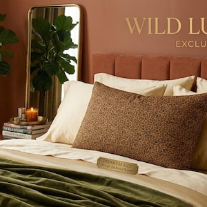 May include: A bedroom scene featuring a bed with cream-colored pillows and a brown leopard-print pillow. A green blanket is draped over the bed. The wall is a warm reddish-brown color, with the words "WILD LUXURY. EXCLUSIVE PATTERN" in gold.