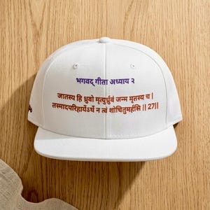 Puede incluir: Gorra de béisbol blanca con texto en sánscrito en rojo y azul. El texto dice "Bhagavad Gita Capítulo 2" y un verso. La gorra está sobre una superficie de madera.