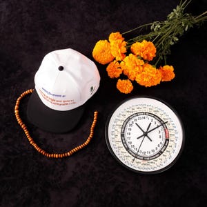 Puede incluir: Una gorra de béisbol blanca con visera negra, texto y un pequeño emblema. Un collar de cuentas de madera, un reloj redondo con manecillas negras y flores naranjas también están presentes.