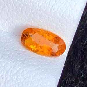 May include: An oval-cut, orange gemstone with multiple facets. The gemstone is set against a white, textured background. The gem's color ranges from light to deep orange, with a brilliant shine.
