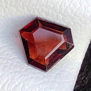 May include: A faceted, reddish-brown gemstone with a unique geometric shape. The gem has a polished surface that reflects light, showcasing its internal structure. The stone is set against a white, textured background, highlighting its color and clarity.