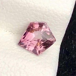 Peut inclure: Une pierre précieuse rose taillée en forme de pentagone. La gemme présente de multiples facettes qui réfléchissent la lumière, créant un effet scintillant. La couleur est un rose clair et doux. La gemme semble être posée sur une surface blanche.