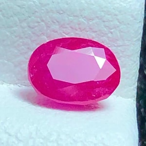 May include: A faceted, oval-shaped ruby gemstone. The gem is a vibrant shade of pinkish-red, with a lighter pink centre. The cut of the stone creates multiple reflective surfaces, enhancing its brilliance. The ruby is set against a white background.