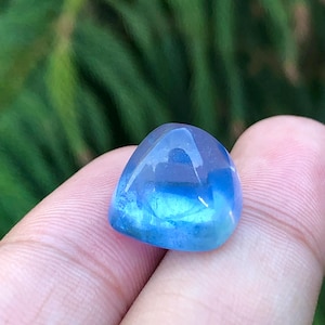 Puede incluir: Una piedra preciosa pulida y translúcida con una forma redondeada e irregular. La piedra pasa de una parte superior clara a un tono azul vibrante en la base. La gema se sostiene entre los dedos sobre un fondo verde borroso.