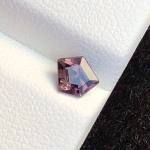 May include: A faceted, purplish-pink gemstone with a unique, geometric cut. The gem is set against a white, textured surface, with a dark background. The stone's facets reflect light, creating a sparkling effect.