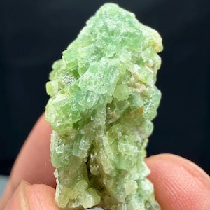 May include: A close-up of a light green mineral specimen. The mineral has a crystalline structure with a slightly rough texture. The color is a vibrant, translucent green, with some areas appearing more opaque. The background is a dark, blurred color.