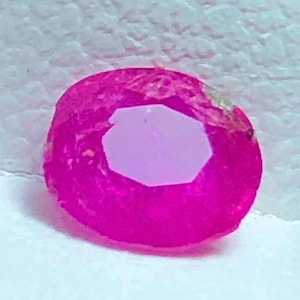 May include: A faceted, oval-shaped gemstone with a vibrant magenta hue. The gem has a lighter, hexagonal-shaped interior. The surface of the gem has a slightly rough texture. The gemstone is set against a white background.