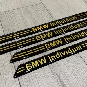 Könnte beinhalten: Vier schwarze Kfz-Zierteile mit goldenen Akzenten und dem Text "BMW Individual". Die Zierteile haben eine glänzende Oberfläche und sind auf einer hellen Holzmaserung angeordnet.
