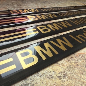 Puede incluir: Primer plano de varios embellecedores automotrices negros con letras doradas que dicen "BMW Individual". Los embellecedores tienen rayas doradas. El fondo es de color neutro.