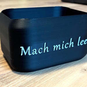 Puede incluir: Un recipiente negro rectangular con esquinas redondeadas. El texto "Mach mich leer!" está impreso en blanco en el lateral. El recipiente parece estar impreso en 3D y tiene un acabado mate. El recipiente está vacío.