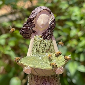 May include: A garden statue of a woman holding a large leaf-shaped bowl, with several bees around it. The statue has a green dress and brown hair. The bowl is light green, and the bees are brown and yellow.