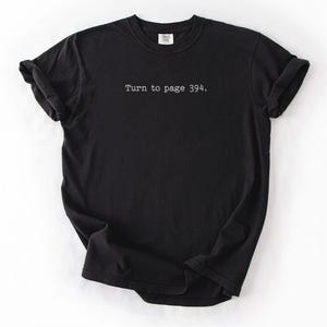 May include: Black t-shirt with the text "Turn to page 394." printed in a simple, sans-serif font. The shirt has a crew neck and short sleeves, with the sleeves rolled up. The fabric appears to be soft and slightly textured.