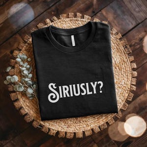 May include: Black t-shirt with the word "Siriusly?" in white, decorative lettering. The shirt is folded on a woven, circular placemat. The background is a wooden surface with some sprigs of greenery.