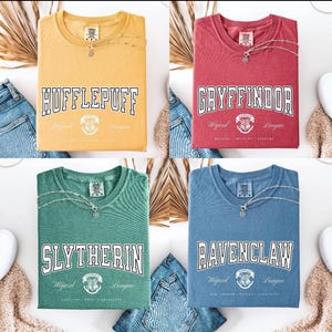 May include: Four short-sleeve t-shirts in yellow, red, green, and blue. Each shirt features a white graphic with the text Hufflepuff, Gryffindor, Slytherin, or Ravenclaw, along with a crest and the words Wizard League printed on the front.