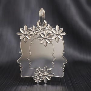 Può includere: Un ciondolo d'argento con due profili di volti adornati da dettagli floreali. Il ciondolo ha un'asola per una catena ed è realizzato in metallo lucido e riflettente. I fiori sono dettagliati e aggiungono un tocco delicato al design.