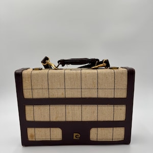 May include: Vintage rectangular suitcase with a tan and brown plaid pattern. The suitcase has a brown leather handle, trim, and gold-colored hardware. The front of the suitcase has the logo of a stylized letter "e" inside a circle.
