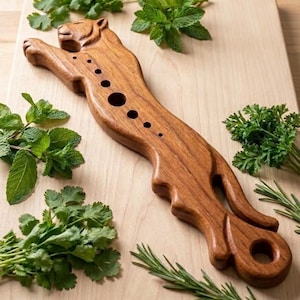 May include: A carved wooden kitchen tool shaped like a leaping tiger, featuring a series of circular holes along its back and a larger circular opening at the tail end, surrounded by fresh sprigs of green herbs on a light wood cutting board.