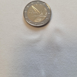 Puede incluir: Una moneda de dos euros con un anillo exterior plateado y un centro dorado. El centro presenta un pájaro estilizado en vuelo, con el texto "EESTI 2023" debajo. La moneda está sobre una superficie blanca.