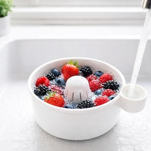 May include: A white bowl containing strawberries, blueberries, and blackberries being washed under a tap. The bowl is in a white sink. The image shows a kitchen setting with a small plant in the background.