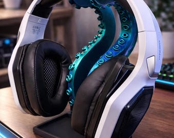 May include: White and black gaming headphones resting on a teal and blue octopus tentacle headphone stand. The headphones have black earcups and a white headband. The stand is on a black base, and the background is blurred.