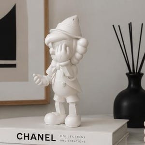 K&ouml;nnte beinhalten: Eine wei&szlig;e Figur einer Zeichentrickfigur mit Hut, Handschuhen und Fliege, die auf einem wei&szlig;en Buch mit dem Aufdruck "CHANEL" steht. Eine schwarze Vase mit Schilf und ein gerahmtes Kunstwerk befinden sich im Hintergrund.