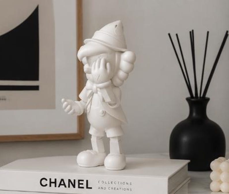 K&ouml;nnte beinhalten: Eine wei&szlig;e Figur einer Zeichentrickfigur mit Hut, Handschuhen und Fliege, die auf einem wei&szlig;en Buch mit dem Aufdruck "CHANEL" steht. Eine schwarze Vase mit Schilf und ein gerahmtes Kunstwerk befinden sich im Hintergrund.