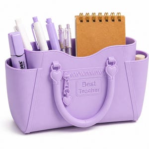 Pode incluir: Um organizador de secretária cor de lavanda em forma de mala com duas pegas, detalhes em forma de coração e um fecho de correr. A parte frontal apresenta o texto "Best Teacher" em relevo num emblema retangular. Contém várias canetas e um caderno.