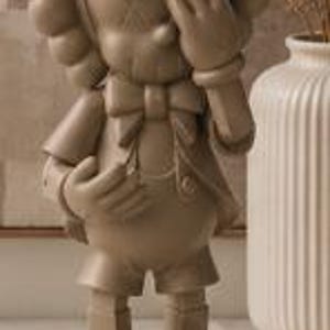 K&ouml;nnte beinhalten: Beige Figur eines Zeichentrickcharakters in klassischer Kleidung, mit Hut und Fliege, eine Hand bedeckt die Augen. Die Figur steht auf einem Buch mit dem Text "at 100 A CENTURY OF STYLE".