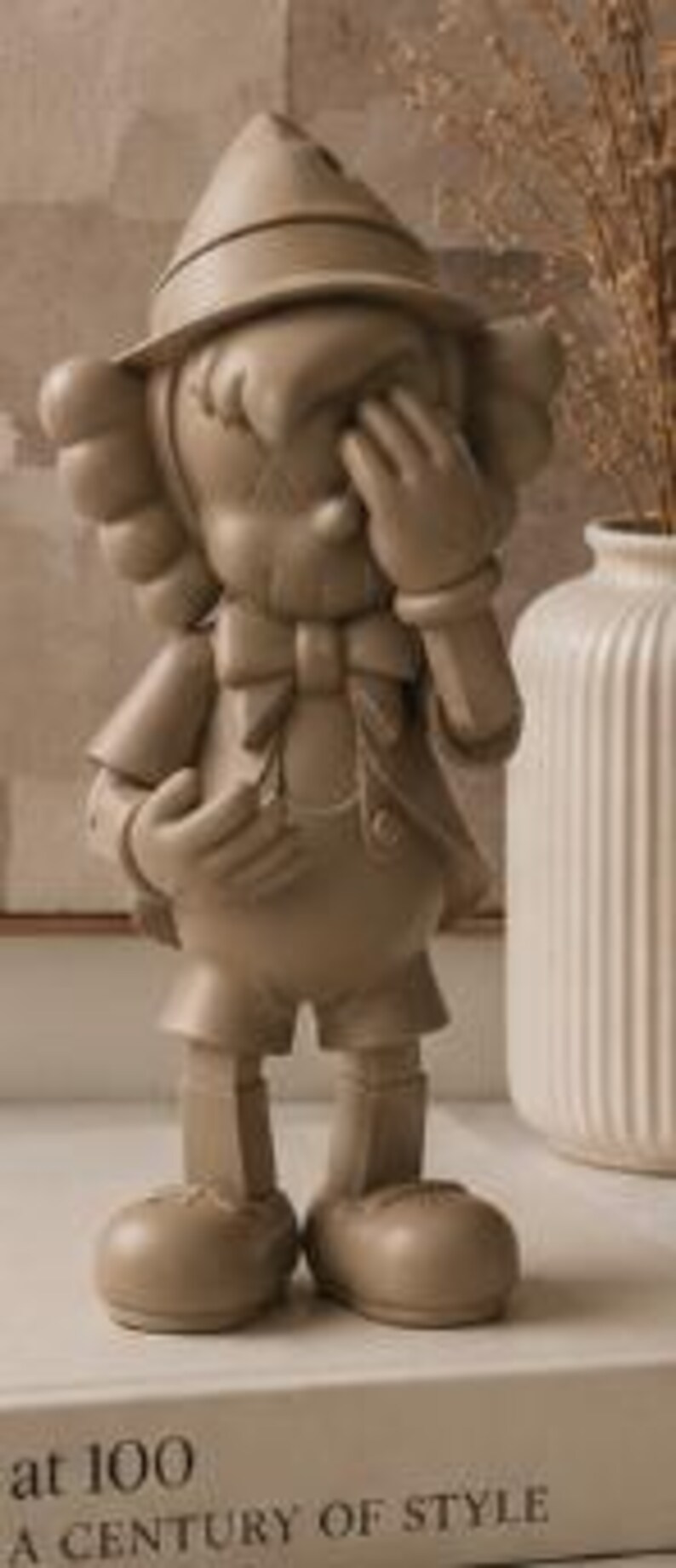 K&ouml;nnte beinhalten: Beige Figur eines Zeichentrickcharakters in klassischer Kleidung, mit Hut und Fliege, eine Hand bedeckt die Augen. Die Figur steht auf einem Buch mit dem Text "at 100 A CENTURY OF STYLE".