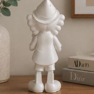 K&ouml;nnte beinhalten: Eine wei&szlig;e Figur einer Zeichentrickfigur, von hinten gesehen. Die Figur tr&auml;gt einen Hut, einen Mantel, Shorts und gro&szlig;e Schuhe. Die Figur steht auf einer Holzoberfl&auml;che, mit einer Vase und B&uuml;chern im Hintergrund.