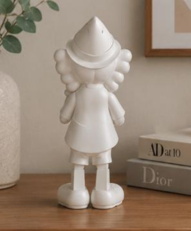 K&ouml;nnte beinhalten: Eine wei&szlig;e Figur einer Zeichentrickfigur, von hinten gesehen. Die Figur tr&auml;gt einen Hut, einen Mantel, Shorts und gro&szlig;e Schuhe. Die Figur steht auf einer Holzoberfl&auml;che, mit einer Vase und B&uuml;chern im Hintergrund.