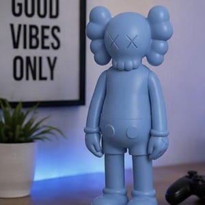 May include: A light blue vinyl figure with crossed-out eyes and a skull-like head. The figure is standing on a wooden surface. A framed print with the words "GOOD VIBES ONLY" is in the background.