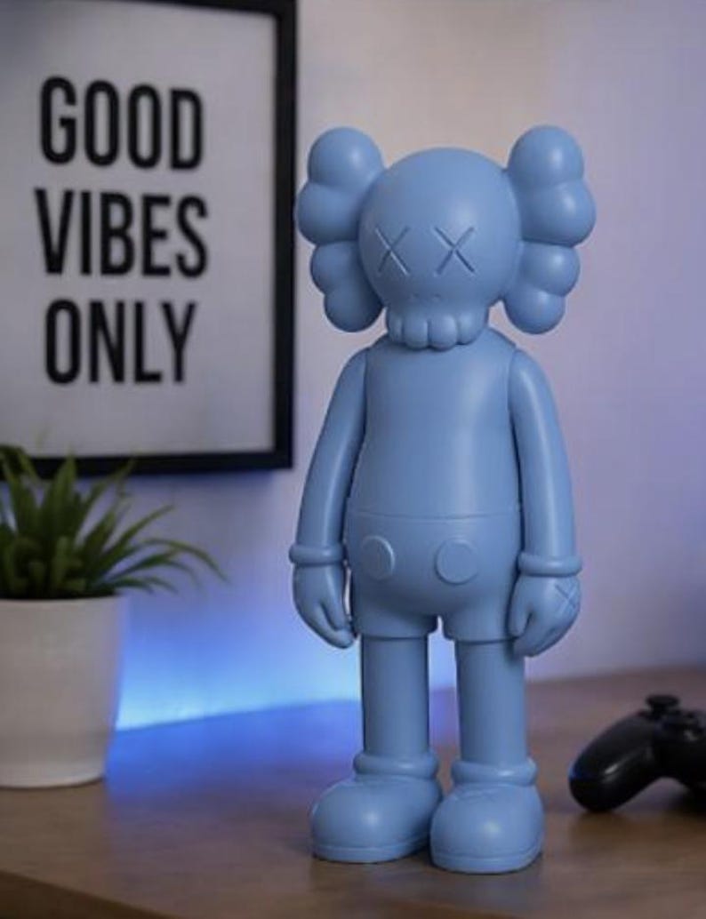 May include: A light blue vinyl figure with crossed-out eyes and a skull-like head. The figure is standing on a wooden surface. A framed print with the words "GOOD VIBES ONLY" is in the background.